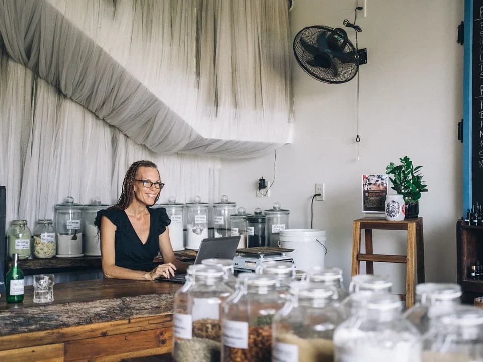 The manager of a bulk-food shop, smiling, working on her laptop behind her counter
